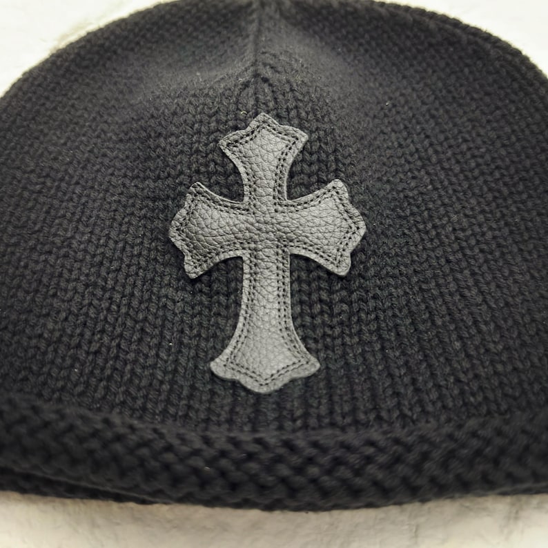 Puede incluir: Gorro de punto negro con una cruz de cuero. El gorro est&aacute; hecho de un material de punto grueso y texturizado. La cruz es de color gris oscuro y tiene una superficie texturizada.