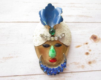 May include: A vintage brooch featuring a stylized face with a white turban, blue and green jewels, and a blue and white feather. The face has a green nose, red lips, and black eyes.
