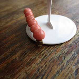 May include: A pink beaded hair clip with a gold metal clasp. The clip is on a white display stand.