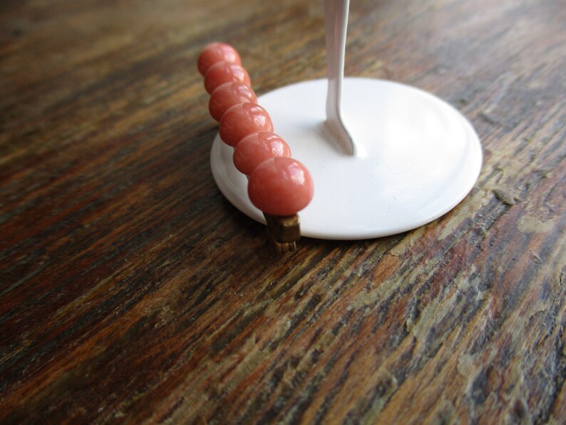May include: A pink beaded hair clip with a gold metal clasp. The clip is on a white display stand.