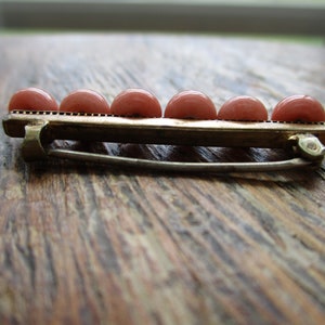 May include: A gold-toned brooch with a row of six pink round stones. The brooch has a safety pin closure.