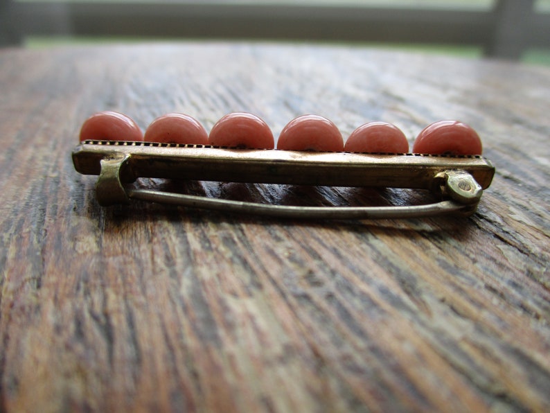 May include: A gold-toned brooch with a row of six pink round stones. The brooch has a safety pin closure.