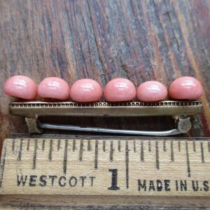 May include: A gold-toned barrette with a row of seven pink cabochon stones. The barrette is about 1 inch long and is sitting on a wooden ruler.