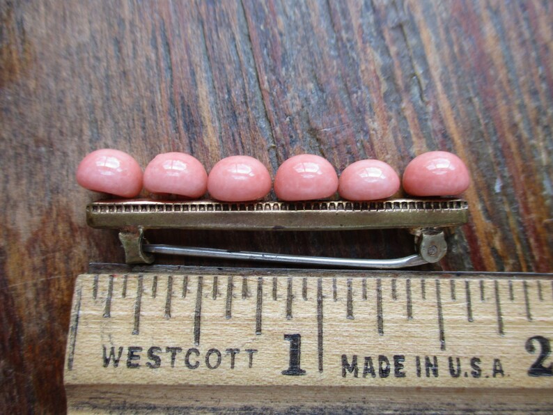 May include: A gold-toned barrette with a row of seven pink cabochon stones. The barrette is about 1 inch long and is sitting on a wooden ruler.