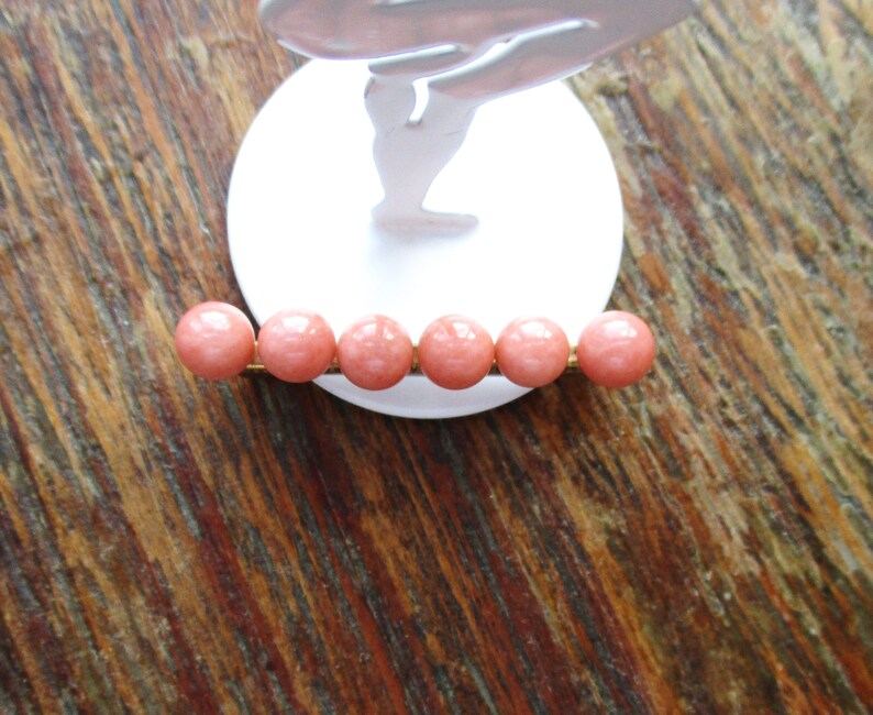May include: A gold bar with six round, pink beads. The beads are evenly spaced on the bar.