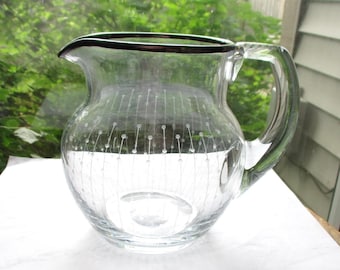May include: Clear glass pitcher with a rounded body and a handle. The pitcher has a dark rim and is decorated with a pattern of vertical lines with small white dots. The pitcher is sitting on a white surface.