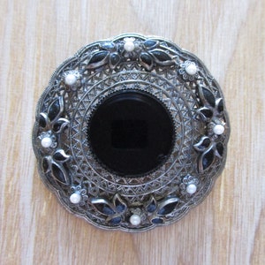 May include: A silver filigree brooch with a black onyx center and white pearl accents.