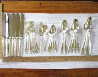 May include: A collection of silver-colored flatware, including knives, forks, spoons, and serving spoons, arranged on a white surface. The silverware has decorative floral detailing on the handles. A wooden ruler is placed along the edge of the flatware.