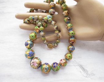 May include: A long necklace made of round, multi-colored beads with floral designs. The beads are made of cloisonné enamel and have gold accents. The necklace is on a hand model.