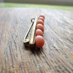 May include: A gold bar pin with a row of seven pink coral beads. The pin is on a wooden surface.
