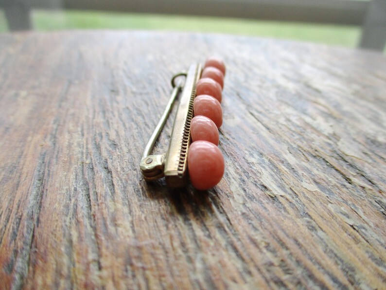 May include: A gold bar pin with a row of seven pink coral beads. The pin is on a wooden surface.