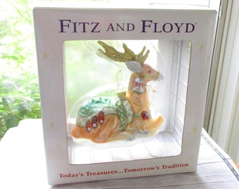 May include: A Fitz and Floyd Christmas ornament of a reindeer, displayed in its original box. The reindeer is tan with gold antlers, a green blanket, and red accents. The box reads "Today's Treasures...Tomorrow's Tradition."