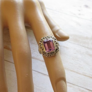 May include: A silver ring with a rectangular, light purple gemstone. The ring is set with small, dark stones around the main stone, creating a decorative border. The ring is on a finger.