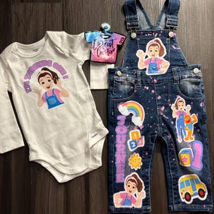 Puede incluir: Conjunto infantil de body blanco de manga larga y peto vaquero. El body dice "I'M TURNING ONE!" y presenta una niña de dibujos animados. El peto tiene un arcoíris, un autobús escolar y otros gráficos.