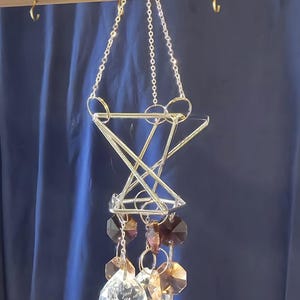 May include: A silver-colored geometric hanging mobile with clear and brown crystal accents. The mobile features a star-like structure, a clear crystal ball, and a crescent moon charm. The mobile hangs from a wooden beam.
