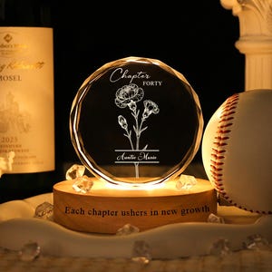 May include: A lit-up, round crystal with a floral design and the text "Chapter Forty" and "Auntie Marie". A baseball and a wine bottle are in the background. The base of the crystal has the text "Each chapter ushers in new growth".