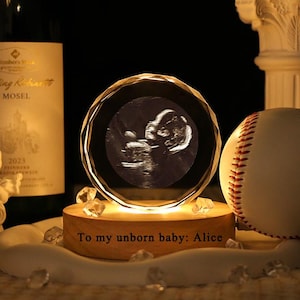Op de afbeelding: Een kristallen bol met een echografie van een baby, verlicht op een houten voet. Een honkbal en een fles wijn staan op de achtergrond. De tekst "To my unborn baby: Alice" staat op de voet.