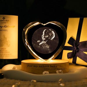 May include: A heart-shaped crystal keepsake with an ultrasound image of a baby, illuminated by a warm light. The base is engraved with the words "For my unborn baby...Love your mother." A bottle of wine and a gift box with a black ribbon are in the background.