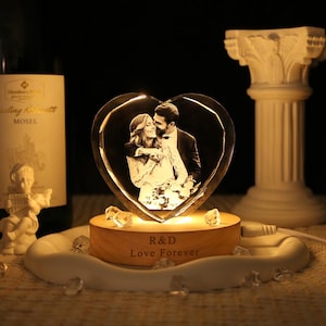 May include: A heart-shaped glass block featuring a laser-etched photograph of a couple, illuminated by a round wooden base. The base is engraved with the text R&D and Love Forever, and is surrounded by small clear crystals on a white tray.