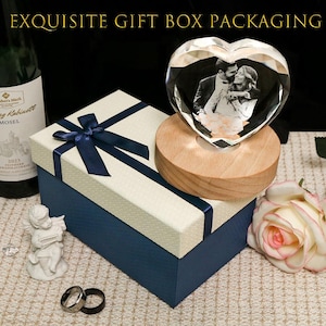 May include: A heart-shaped crystal block featuring a black and white photograph of a couple, mounted on a round wooden base. The set includes a cream and navy blue gift box with a ribbon bow, a wine bottle, a small cherub figurine, and two rings.