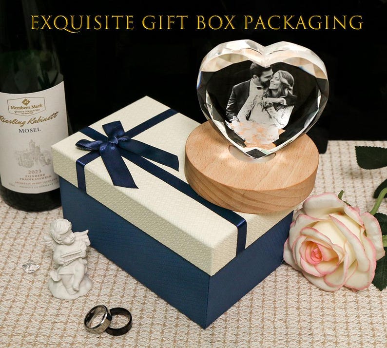 May include: A heart-shaped crystal block featuring a black and white photograph of a couple, mounted on a round wooden base. The set includes a cream and navy blue gift box with a ribbon bow, a wine bottle, a small cherub figurine, and two rings.