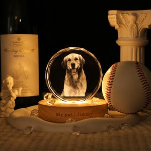 May include: A circular crystal plaque featuring a laser-etched portrait of a golden retriever, mounted on a round wooden base illuminated from below. The base is engraved with the text "My pet : Jimmy" and sits near a baseball and wine bottle.