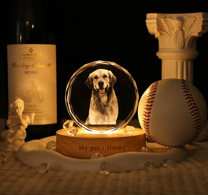 May include: A circular crystal plaque featuring a laser-etched portrait of a golden retriever, mounted on a round wooden base illuminated from below. The base is engraved with the text "My pet : Jimmy" and sits near a baseball and wine bottle.