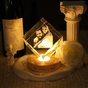 Puede incluir: Un cubo de fotos de cristal con una foto de un hombre y una niña, iluminado por una luz cálida. El cubo se encuentra sobre una base de madera con la inscripción "My favorite dad". Una rosa blanca y una botella de vino en el fondo.
