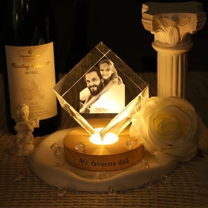 May include: A diamond-shaped glass block featuring a laser-etched photograph of a man and a child, resting on a circular wooden base illuminated from below. The base is inscribed with the text My favorite dad in a dark, sans-serif font.