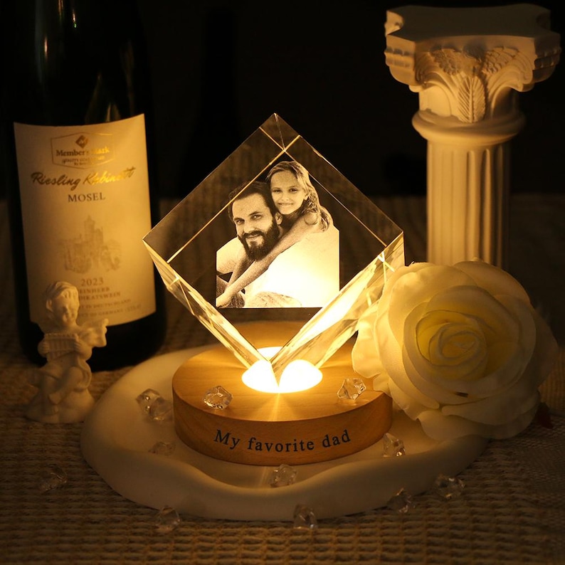 May include: A diamond-shaped glass block featuring a laser-etched photograph of a man and a child, resting on a circular wooden base illuminated from below. The base is inscribed with the text My favorite dad in a dark, sans-serif font.