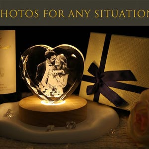 May include: A heart-shaped glass block featuring a laser-etched photograph of a couple, illuminated by a round wooden light base. The text "PHOTOS FOR ANY SITUATION" appears at the top. A gift box with a dark blue ribbon sits to the right.