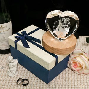 May include: A heart-shaped crystal photo block with a couple's image, set on a wooden base, next to a blue gift box with a navy ribbon. A white and pink rose and a small angel figurine are also present.