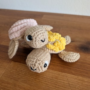 Op de afbeelding: Een handgemaakte gehaakte schildpad in beige, roze en geel. De schildpad heeft een beige kop en lichaam, een roze schild en gele accenten. Het heeft zwart-witte ogen en een lachende uitdrukking. Een zacht, handgemaakt speelgoed.