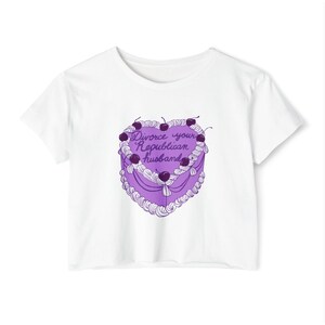 May include: White t-shirt featuring a heart-shaped cake design in purple with cherries on top. The cake has the text "Divorce your Republican husband" in a cursive font. The shirt is a cropped style.