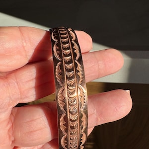 Może przedstawiać: Miedziana bransoletka z misternymi, wytłoczonymi wzorami. Wzór składa się z powtarzających się geometrycznych kształtów i linii, tworząc teksturowaną powierzchnię. Bransoletka jest trzymana w dłoni, ukazując jej wypolerowane wykończenie i szczegółowe rzemiosło.