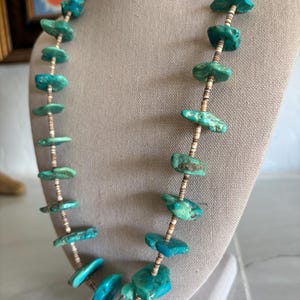 Puede incluir: Collar de piedras turquesas con forma orgánica irregular. El collar presenta piedras de turquesa pulidas ensartadas en un cordón beige. Las piedras varían en tonos azules y verdes, creando un aspecto único y artesanal.