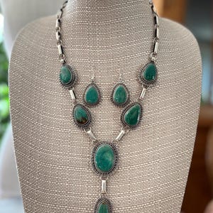 May include: A jewelry set consisting of a necklace and matching drop earrings. The necklace features a silver chain with multiple teardrop and oval green stones set in decorative silver frames. The earrings mirror the teardrop stone design.