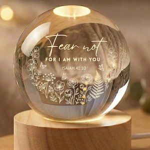 Op de afbeelding: Een heldere glazen bol met de woorden "Fear not, for I am with you" en bloemmotieven die erin zijn geëtst. De bol staat op een ronde houten basis, van binnenuit verlicht. De tekst komt uit Jesaja 41:10.