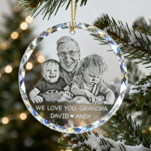 Op de afbeelding: Een heldere, ronde kerstversiering met een familiefoto. De ornament heeft een facetrand en een gouden draad om op te hangen. De foto toont een opa met twee jonge kinderen, met de tekst "WE LOVE YOU, GRANDPA DAVID ANDY".