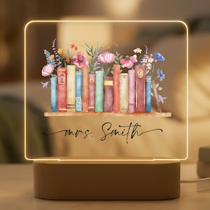 May include: A personalized night light featuring a watercolor illustration of books and flowers. The design includes a wooden shelf, colorful books, and the name "mrs. Smith" in elegant script. The light has a warm glow.