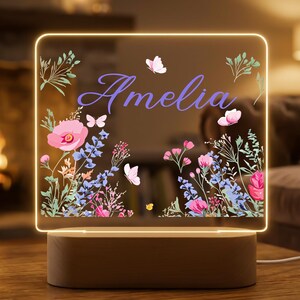 Puede incluir: Una luz nocturna LED personalizada con el nombre "Amelia" en escritura púrpura. El panel acrílico presenta un diseño floral con amapolas rosas, flores azules y mariposas. La luz se asienta sobre una base de madera.