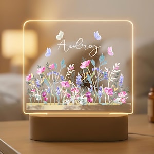 Puede incluir: Una luz de noche personalizada con un diseño floral. El panel acrílico presenta flores rosas, moradas y azules, mariposas y el nombre "Aubrey" en cursiva. La luz se asienta sobre una base beige.