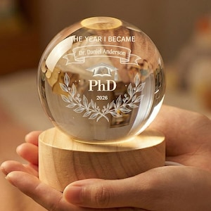Puede incluir: Una esfera de vidrio transparente grabada con "THE YEAR I BECAME" y "PhD 2026" se asienta sobre una base de madera. La esfera tiene una pancarta con el nombre "Dr. Daniel Anderson" y un diseño de corona de laurel. La base está iluminada desde dentro.