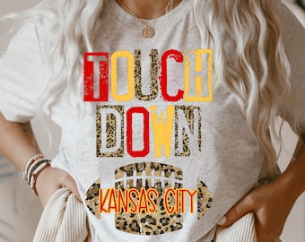 May include: A gray t-shirt with the words "TOUCHDOWN" in red, yellow, and black letters with a leopard print pattern. Below the words is a football with the words "KANSAS CITY" inside.