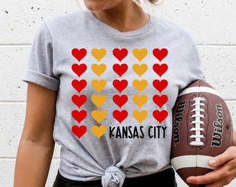May include: Gray t-shirt with a pattern of red and yellow hearts. The text "KANSAS CITY" is printed below the hearts. A person is holding a brown football.
