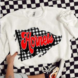 May include: White crew neck sweatshirt with a red and black chequered flag design and the word "Hornets" in red script. The sweatshirt is displayed on a black and white chequered surface.