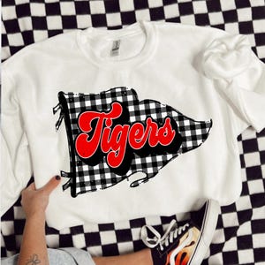 May include: White crew neck sweatshirt with a black and white checked flag design and the word "Tigers" in red script. The sweatshirt is displayed on a black and white checked surface.
