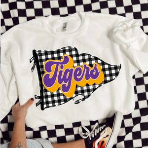 May include: White sweatshirt with a graphic of a flag displaying the word "Tigers" in purple and yellow. The flag design has a black and white checkered pattern. The sweatshirt is shown on a checkered surface.