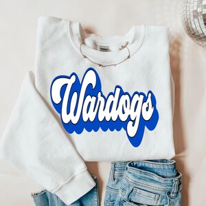 May include: A white sweatshirt with the word "Wardogs" in a blue and white retro font. The sweatshirt is folded with a pair of blue jeans. A delicate gold necklace is draped across the sweatshirt.