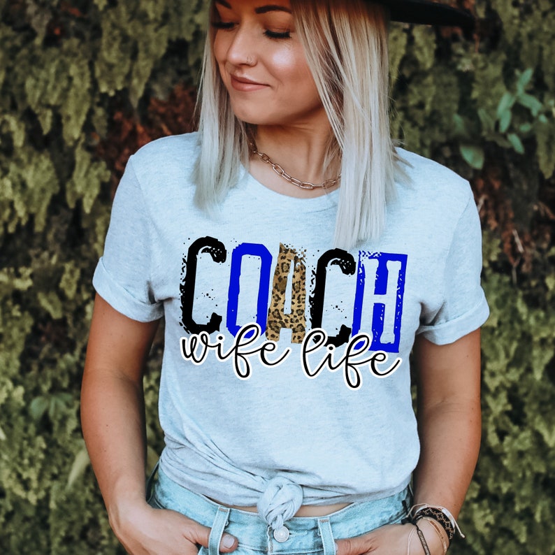 Może przedstawiać: Jasnoniebieska koszulka z napisem "COACH wife life" w kolorze czarnym, niebieskim i w nadruku w panterkę.